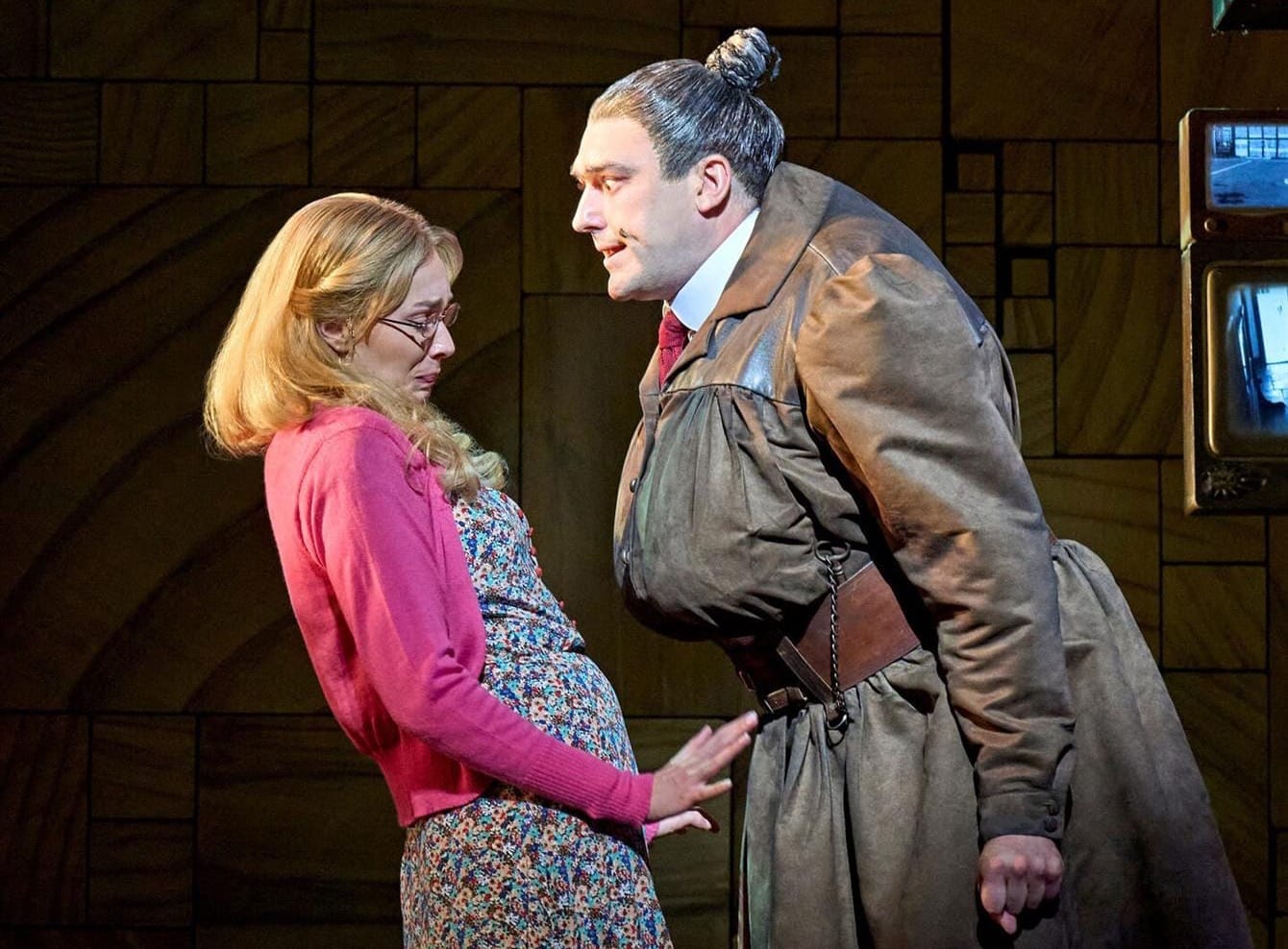 The terrifying woman massive woman with a tight grey bun wearing military-inspired garb leans menacingly over a small scared woman in a floral dress and pink cardigan.