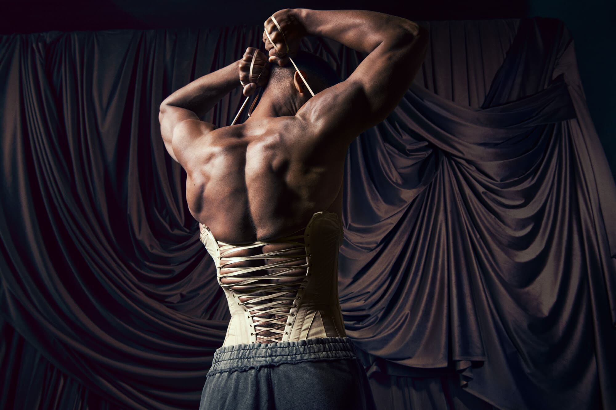 A muscular cis man is pictured from the back, attempting to lace his muscular torso into a satin corset.