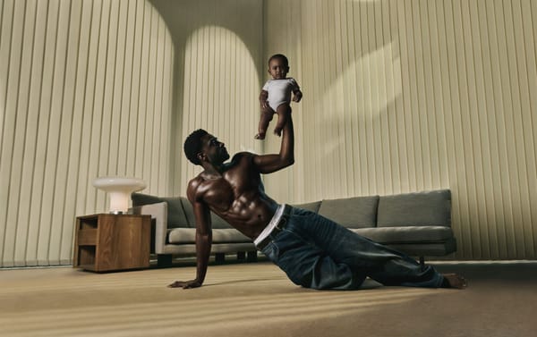 A shirtless, muscular man poses on the floor of a stylish living room. He uses one arm to prop himself up and the other to hoist an adorable baby up high in the air.
